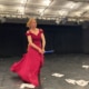 Frau in rotem Kleid in einem schwarzen Theaterraum "reitet" auf einem Bestenstiel durch den Raum