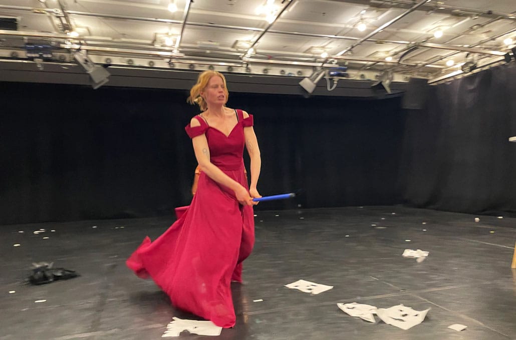 IFANT-Werkraum-Theater_Beitragsbild Frau in rotem Kleid in einem schwarzen Theaterraum "reitet" auf einem Bestenstiel durch den Raum