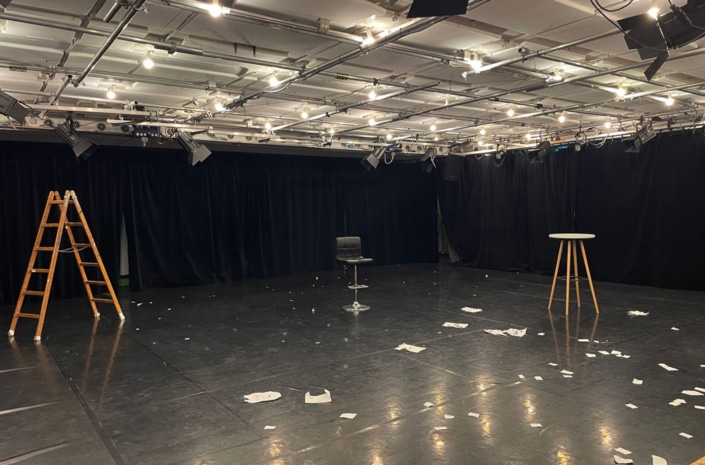 IFANT_Werkraum-Theater03 Schwarzer Theaterraum in dem eine Leiter, ein Barstuhl zum Drehen und ein dreibeiniger Stehtisch stehen. auf dem Boden liegen viele Papierschnipsel